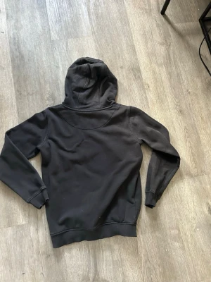 Svart hoodie från Lyle & Scott - Säljer en svart hoodie från Lyle & Scott med klassisk gul logga på bröstet. Hoodien har huva, magficka och ribbade muddar vid ärmar och nederkant. Perfekt för en avslappnad och stilren look. Materialet känns mjukt och bekvämt.