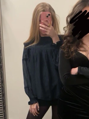 Mörkblå vid blus med volang - Snygg mörkblå blus med lös passform och volangdetalj över bröstet. Blusen har långa, transparenta ärmar med resår vid handlederna och hög hals. Perfekt för en trendig och avslappnad look.