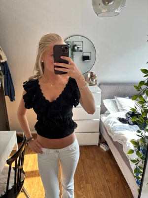 Svart volangtopp med v-ringning - Supersöt svart topp med djup v-ringning och volangdetaljer längs axlar och byst. Croppad modell med resår i midjan och korta ärmar. Perfekt för dig som vill ha en trendig och feminin look.