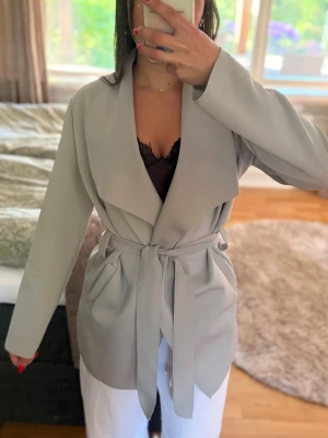 Ljusgrå trenchcoat med knytskärp - Stilren ljusgrå trenchcoat med bred krage och knytskärp i midjan. Jackan har en avslappnad passform och två stora fickor framtill. Perfekt för dig som gillar minimalistisk och modern stil. Materialet ser ut att vara mjukt och följsamt.