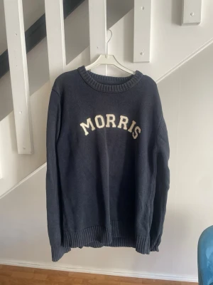 Mörkblå tröja från Morris - Mörkblå stickad tröja från Morris med vit text på bröstet. Tröjan har rund halsringning och ribbade muddar vid ärmslut och nederkant. Perfekt för en avslappnad och stilren look.