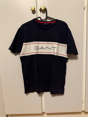 GANT t-shirt  - Snygg svart t-shirt från GANT med stor vit panel och röd detalj över bröstet där loggan är tryckt. Klassisk rund hals och korta ärmar. Tillverkad i mjuk bomull som känns skön mot huden. Perfekt till jeans eller shorts för en clean look.