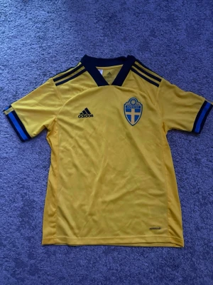 Svensk landslagströja Adidas gul - Gul svensk landslagströja från Adidas med blå detaljer och svart logga. Tröjan har korta ärmar, V-ringad krage och Sveriges landslagsemblem på bröstet. Tillverkad i lätt och ventilerande material, perfekt för fotboll eller träning eller för att heja fram Sverige i sommar när vi ska gräva guld. Inte använd en enda gång då den var för liten och nu bara legat i en låda.