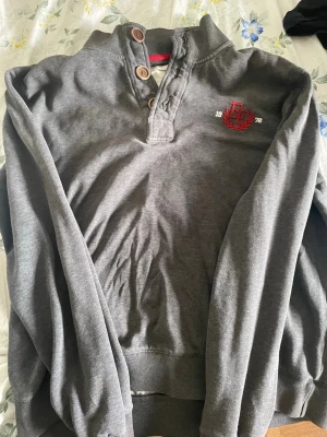 Grå sweatshirt med brodyr och knappar - Mysig grå sweatshirt med långa ärmar och ribbade muddar. Tröjan har en knappslå med två träknappar vid halsen och en broderad detalj på bröstet med röd logga och texten 1972. Perfekt för en avslappnad stil.