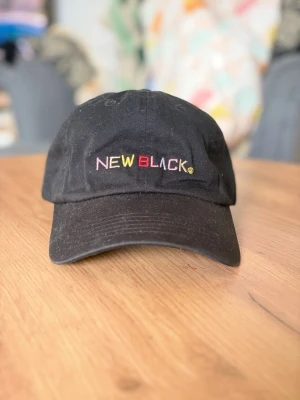 Svart keps från New Black - Säljer en svart keps från New Black med broderad logga i flera färger framtill. Klassisk böjd skärm och justerbart spänne bak för perfekt passform. Tillverkad i bomullstyg som andas. Perfekt accessoar för en avslappnad streetwear-look.