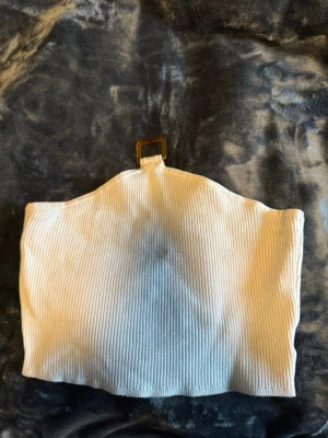 Vit ribbad bandeau topp - Säljer en vit ribbad bandeau topp med en snygg detalj i form av ett fyrkantigt spänne framtill. Toppen är axelbandslös och har en croppad passform som passar perfekt till höga jeans eller kjol.