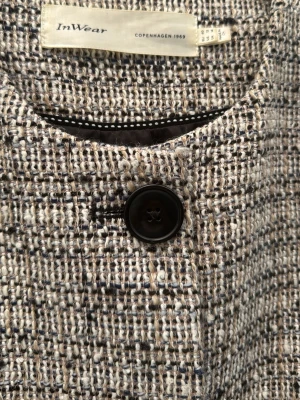 Tweedkavaj från InWear med stora knappar - Snygg tweedkavaj från InWear i beige, svart och grå toner. Rak, boxig passform med stora svarta knappar framtill och två fickor med lock. Materialet är grovt vävt och ger en klassisk, trendig look. Perfekt för dig som gillar stilrena detaljer och modern design.