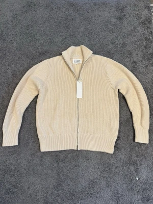 Beige stickad ziptröja Maison Margiela - Säljer en beige stickad ziptröja från Maison Margiela. Tröjan har hög krage, grovstickad struktur och dragkedja framtill. Perfekt för lager på lager och har en clean, minimalistisk look. Materialet känns mjukt och exklusivt.