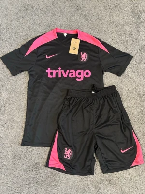 Chelsea träningsset Nike svart/rosa - Chelsea FC träningsset från Nike i svart med rosa detaljer. Setet består av en kortärmad tröja och shorts, båda med klubbmärke och Nike-logga. Materialet är lätt och andas, perfekt för fotbollsträning eller gym. Trivago-logga på bröstet.