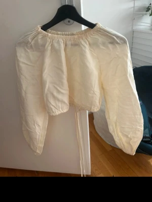 Krämvit blus med puffärmar - Trendig krämvit blus med puffiga långärmar och resår vid halsringningen. Blusen har en avslappnad passform och knytband i midjan för en justerbar look. 