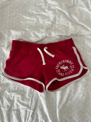 Röda shorts från Abercrombie & Fitch - Snygga röda shorts från Abercrombie & Fitch med vit snörning och vita kanter. Tryck med logga och älg på ena benet. Perfekta för sommaren och chill dagar. Mjukt material och sportig look.