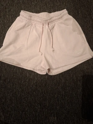 Ljusrosa shorts med Los Angeles-tryck - Säljer ett par ljusrosa mjukisshorts med resår i midjan och dragsko. Shortsen har sidofickor och ett diskret 'Los Angeles'-tryck på ena benet. Perfekta för chill dagar eller träning. Materialet känns mjukt och bekvämt, troligen bomull eller bomullsblandning.