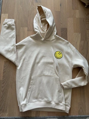 Beige hoodie med smileytryck - Snygg beige hoodie med huva och känguruficka framtill. På bröstet finns ett gult smileytryck som ger en cool och avslappnad vibe. Perfekt för dig som gillar streetwear och vill ha något bekvämt och stilrent. Materialet känns mjukt och skönt mot huden.