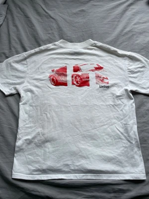 Vit Carhartt t-shirt med biltryck - Vit t-shirt från Carhartt med ett litet svart tryck fram och ett stort rött bilmotiv samt texten 'Drive' på ryggen. Klassisk passform och rund halsringning. 