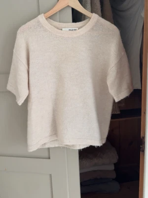 Beige stickad t-shirt från Selected Femme - Kortärmad stickad t-shirt i mjuk beige ullmix från Selected Femme. Tröjan har rund halsringning och avslappnad passform, perfekt för lager-på-lager och en chill stil.