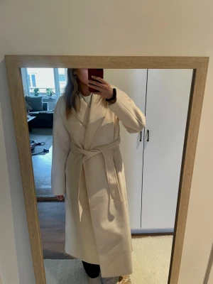 Lång kappa Gina Tricot  - Créme färgad lång kappa från Gina Tricot. Helt ny med lapparna kvar. Storlek xs med skulle säga att den sitter som en s. Har en jätte liten fläck (se sista bilden) men som lätt går att tvätta bort. Köpt för 900kr