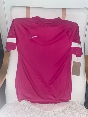 Rosa sporttröja från Nike - Snygg rosa sporttröja från Nike med vit logga på bröstet och vita detaljer på ärmarna. Tillverkad i ett lätt och snabbtorkande material som passar perfekt till träning eller match. Klassisk rund hals och korta ärmar för maximal rörelsefrihet. Storlek S. Den är lite klippt där nere 