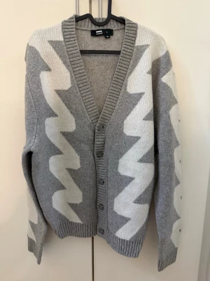 Grå kofta med vitt zigzag-mönster - Säljer en grå kofta från Weekday med vitt zigzag-mönster. Koftan har djup V-ringning, ribbstickade muddar och knäppning framtill med fyra knappar. Perfekt för lager-på-lager och har en avslappnad passform.