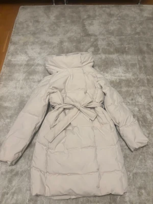 Ljusbeige pufferjacka med knytskärp -  Ljusbeige pufferjacka med hög krage och knytskärp i midjan. Jackan har quiltad design och långa ärmar.Stängs med dragkedja och tryckknappar framtill. 