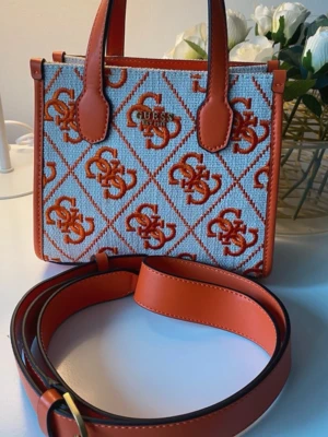 Guess Silvana mini handväska orange - Säljer en Guess Silvana 2 Compartment Mini handväska i orange och beige med mönstrat tyg och logotypdetaljer. Väskan har både handtag och en bred axelrem i orange skinnimitation. Snygg design med GG-mönster och metallogo framtill. Använd endast 1 gång nypris 1295:-