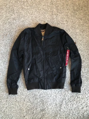 Svart bomberjacka Alpha Industries - Klassisk svart bomberjacka från Alpha Industries med orange foder och ribbade muddar. Jackan har dragkedja framtill, två fickor med tryckknapp och ikonisk röd tag på ärmen. Perfekt för en cool streetstil och tidlös look.