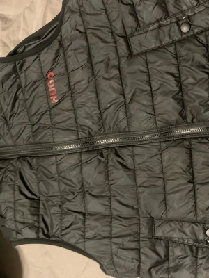 Svart dunväst från Hugo boss  - Snygg svart dunväst från Hugo med quiltat mönster och diskret röd logga på bröstet. Västen har dragkedja framtill och praktiska fickor med knappstängning. Perfekt för lager på lager och ger en clean look till din outfit.