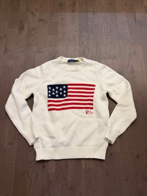 Vit stickad tröja Polo Ralph Lauren - Vit stickad tröja från Polo Ralph Lauren med amerikansk flagga i rött, vitt och blått på bröstet samt RL-broderi. Tröjan har rund halsringning och ribbstickade muddar vid ärmslut och nederkant. Perfekt för dig som gillar klassisk preppy stil.