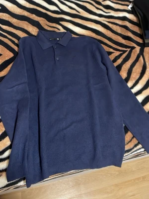 Mörkblå pikétröja med lång ärm - Stilren mörkblå pikétröja med lång ärm och klassisk krage. Tröjan har två knappar vid halsen och är tillverkad i ett mjukt, tunt material som passar perfekt till en avslappnad men ändå uppklädd look.