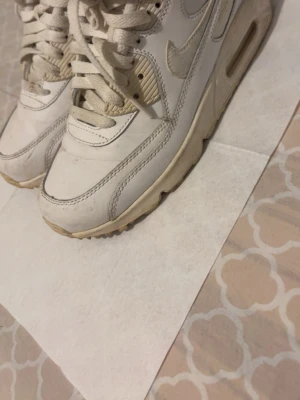 Vita Nike Air Max 90 sneakers - Säljer ett par klassiska vita Nike Air Max 90 sneakers med beige detaljer. Skorna har en chunky sula, synlig Air-enhet i hälen och är gjorda i en mix av läder och mesh. Perfekta för dig som gillar retrostil och streetwear.