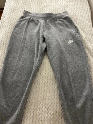 Gråa mjukisbyxor från Nike - Säljer ett par klassiska gråa mjukisbyxor från Nike med broderad logga på vänster ben. Byxorna har resår i midjan och vid bensluten samt sidofickor. Perfekta för chill dagar eller träning. Materialet är mjukt och bekvämt, troligtvis bomullsblandning.