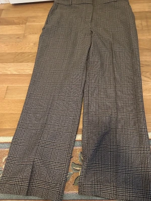 Rutiga kostymbyxor i brunt och svart - Snygga kostymbyxor med klassiskt rutigt mönster i bruna och svarta toner. Byxorna har raka ben och pressveck för en stilren look. Perfekta för dig som gillar en tidlös och trendig stil.