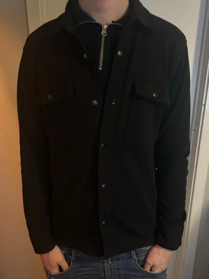 Svart overshirt med fickor - Stilren svart overshirt med klassisk krage och tryckknappar framtill. Perfekt för hösten. Funkar med både hoodie, skjorta och t shirt. Köpt för 1200kr ifrån selected homme. Gott skick. Pris går att diskutera.