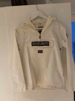 Vit vindjacka från Napapijri - Snygg vit vindjacka från Napapijri med huva och dragkedja vid halsen. Stor ficka framtill med Napapijri-logga och norsk flagga. Jackan har elastiska muddar och justerbar nederkant. Perfekt för blåsiga dagar och enkel att matcha.