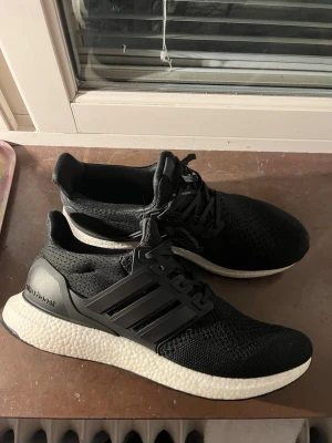 Adidas Ultraboost svarta sneakers - Svarta Adidas Ultraboost sneakers med stickad ovandel och klassiska tre ränder på sidan. Skorna har snörning och en vit, responsiv Boost-sula som ger grym komfort. Perfekta för dig som gillar stilrena och sportiga skor med modern känsla.