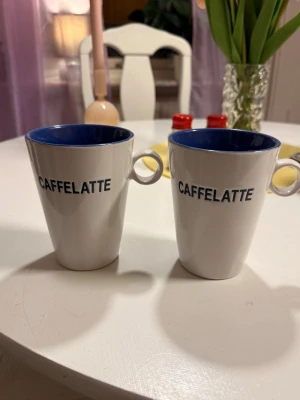 Två vita caffelatte-muggar - Säljer två stilrena vita muggar med blå insida och texten 'CAFFELATTE' i svart på framsidan. Perfekta för kaffeälskare som vill ha en snygg kopp till morgonkaffet. Muggarna har rund form och praktiskt handtag.