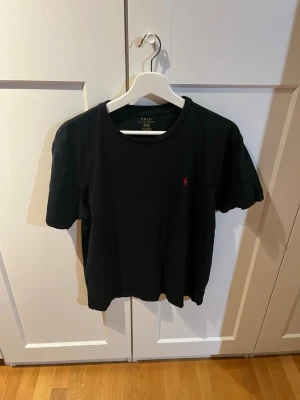 Svart t-shirt från Polo Ralph Lauren - Svart t-shirt från Polo Ralph Lauren med klassisk passform och liten röd logga broderad på bröstet. T-shirten har rund halsringning och är tillverkad i mjuk bomull. Perfekt basplagg för en clean och stilren look.