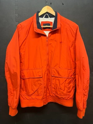 Röd bomberjacka från Lacoste - Snygg röd bomberjacka från Lacoste med dragkedja framtill, två stora fickor med lock och knapp, samt ribbade muddar vid ärmslut och midja. Jackan har en klassisk krage med mörkblå detalj och är tillverkad i ett lätt, glansigt material.