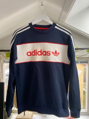 Mörkblå Adidas sweatshirt - Snygg mörkblå sweatshirt från Adidas med klassiska vita ränder på axlarna och stort vitt fält med röd Adidas-logga och treklöver på bröstet. Tröjan har rund halsringning och ribbade muddar. Perfekt för en sportig och avslappnad stil.