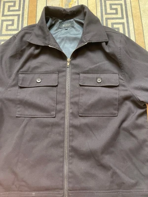 Svart overshirt från Riley med dragkedja - Stilren svart overshirt från Riley med klassisk krage, två stora bröstfickor med lock och knapp samt dragkedja framtill. Overshirten har raka ärmar och är tillverkad i ett slätt tyg som ger en clean look. Perfekt för lager-på-lager-stil. Den är i jätte bra skick frågor och funderingar skriv gärna. Priset går och diskuteras vid snabbaffär. 