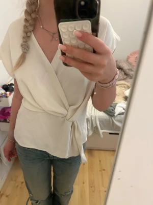 Vit omlottopp med knyt - Säljer en vit omlottopp med knyt i midjan och v-ringad hals. Toppen har korta ärmar och en avslappnad passform, perfekt till jeans eller kjol. Liten fläck där fram men man märker inte av den