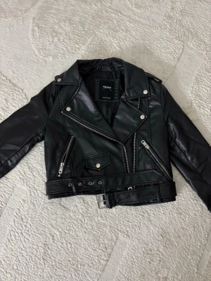 Svart skinnjacka från H&M Divided - Snygg svart skinnjacka från Pull & Bear med klassisk bikerstil, silvriga dragkedjor, tryckknappar och bälte i midjan. Jackan har flera fickor och coola detaljer som ger en edgy look. Perfekt för dig som vill ha en trendig och tidlös jacka.