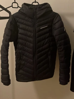 Svart dunjacka från Peak Performance - Snygg svart dunjacka från Peak Performance med huva och dragkedja framtill. Jackan har quiltad design och logga på bröstet. Perfekt för kalla dagar och enkel att matcha med olika outfits.