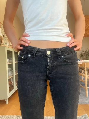 Svarta lågmidjade bootcut jeans med silverknappar - Snygga svarta lågmidjade jeans med bootcut ben och klassiska fem fickor. Jeansen har silverfärgade knappar samt extra detaljer på fickorna bak. Perfekta till en enkel t-shirt eller hoodie. Materialet är denim med lite stretch för skön passform. Follarna där nere är uppsprättade 
