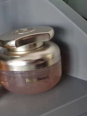 Rare Beauty parfym guldrosa - Lyxig parfym från Rare Beauty i en rund flaska med guldig lock och rosa glas. Flaskan har en modern och elegant design med metallisk finish och tydlig logga på framsidan. Perfekt för dig som gillar trendiga och snygga dofter.