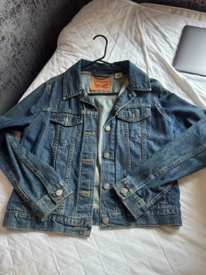Klassisk blå Levi's jeansjacka - Snygg blå jeansjacka från Levi's med klassisk passform, bröstfickor med lock och knappar framtill. Jackan har tydliga sömmar och silverfärgade knappar med Levi's-logga. Perfekt för en cool och avslappnad stil.