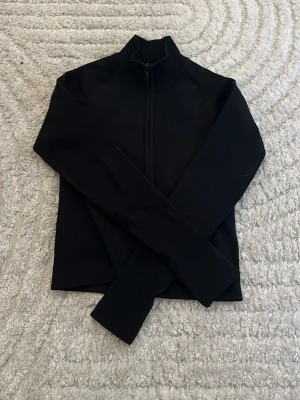 Svart träningskofta från 157 - Snygg svart ziptröja från 157 med hög krage och hel dragkedja framtill. Tröjan har långa ärmar och är tillverkad i ett mjukt och stretchigt material som passar perfekt till lager-på-lager. Enkel och stilren design utan synliga loggor på framsidan.