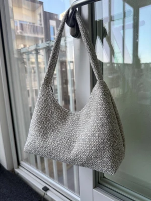 Naturvit axelväska i möbeltyg - Säljer en stilren handväska i naturvit möbeltyg med mjuk struktur. Väskan har en enkel axelrem och stängs med tryckknapp. Insidan är fodrad i mörkbrun tyg. Perfekt för dig som gillar minimalistisk och trendig design. Mått ca 26x15cm