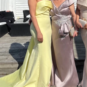 Ljusgul axelbandslös långklänning - Superfin ljusgul långklänning med axelbandslös design och elegant drapering över bysten. Klänningen har en figurnära passform och ett långt släp baktill som ger en riktigt lyxig känsla. Perfekt för dig som vill sticka ut med en fräsch och somrig look. Den är i storlek 38 men har minakat den till 36 men det går att sprätta upp. Nypris:3800kr 