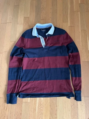 Randig pikétröja från bondelid  - Randig långärmad pikétröja från bondelid i marinblått och vinrött med breda ränder. Klassisk vit krage och knappslå framtill. Perfekt för en avslappnad och vintage stil!