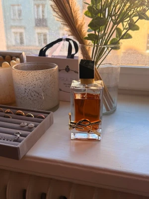 Yves Saint Laurent Libre parfym - Säljer en elegant YSL Libre parfym i en genomskinlig glasflaska med guldiga detaljer och svart asymmetrisk kork. Parfymen har en varm bärnstensfärgad vätska och flaskan pryds av YSL:s ikoniska logga i metall.